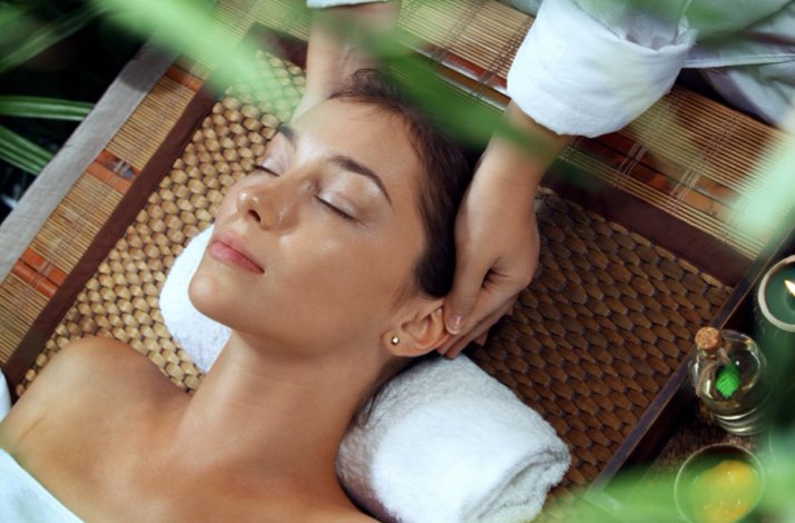 Therapist performing a calming head massage on a woman resting on a bamboo mat in a natural setting. Confidence: High