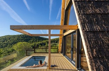 Woman relaxing in a private plunge pool on a cabin deck, overlooking rolling green hills under a blue sky.