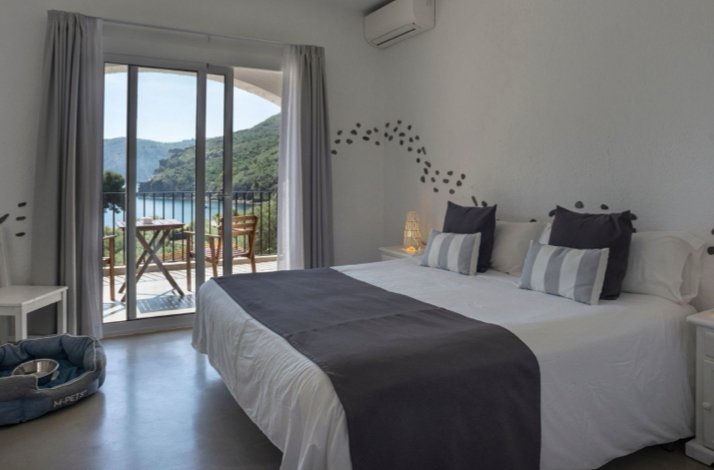 Bright double room at Hotel Restaurante Cala Joncols with grey-and-white bedding and a balcony overlooking the bay.