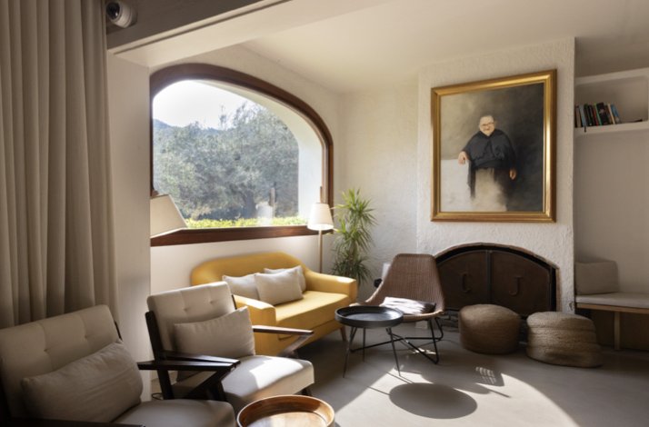 Cosy lounge at Hotel Restaurante Cala Joncols with arched window, yellow sofa, and a framed portrait above the fireplace.
