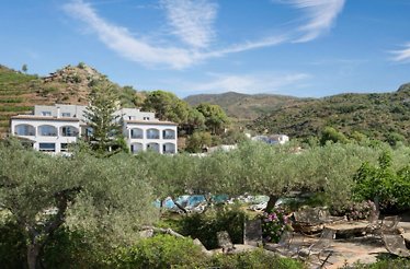 Hotel Restaurante Cala Joncols' white multi-storey building nestled among olive trees, gardens, and terraced hillside.