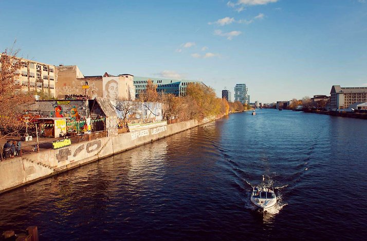 A boat sailing down a river with buildings adorned with street art along the shore.