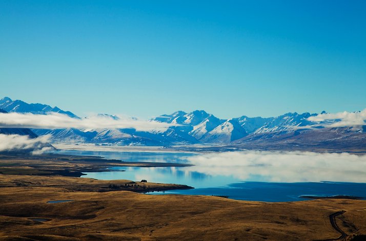 Low clouds hover over the blue waters of Lake Tekapo and the snow-capped Southern Alps in New Zealand.