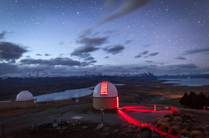Astronomical domes at Mount John Observatory overlook a dark Lake Tekapo under a starry sky in New Zealand.