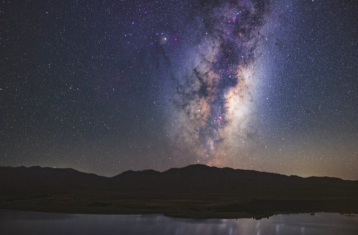 The Milky Way galaxy shines brightly over the dark silhouette of the Aoraki Mackenzie mountains and lake in New Zealand.