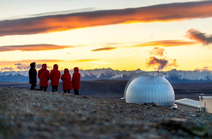 Tourists in red coats look at the Southern Alps and a dome at Mount John Observatory during sunset in New Zealand.