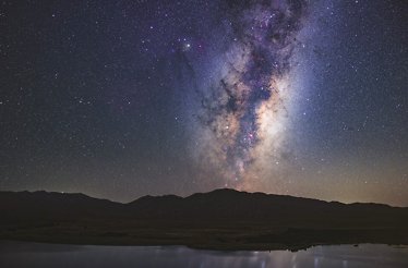 The Milky Way galaxy shines brightly over the dark silhouette of the Aoraki Mackenzie mountains and lake in New Zealand.