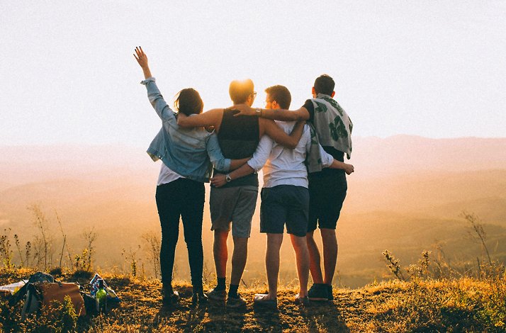Four people stand side by side with their arms around each other, facing a scenic sunset over distant hills in South Africa.