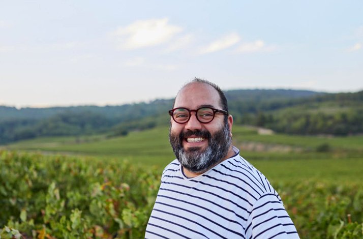 A smiling man wearing glasses standing in a vineyard with rolling green hills in the background.