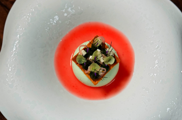 Artfully plated fine-dining dish with herb broth, edible flowers, and golden puree on a sculpted white plate.