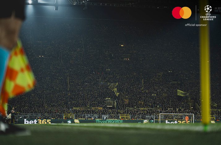 A stand full of football fans watching an UEFA Champions League match. Mastercard and UEFA logos at the top right corner.