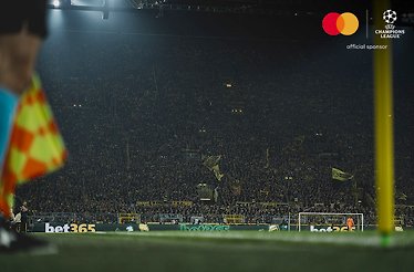 A stand full of football fans watching an UEFA Champions League match. Mastercard and UEFA logos at the top right corner.