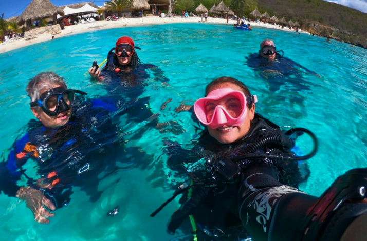 Four scuba divers float together in bright turquoise water near a sandy beach with thatched huts in the background.