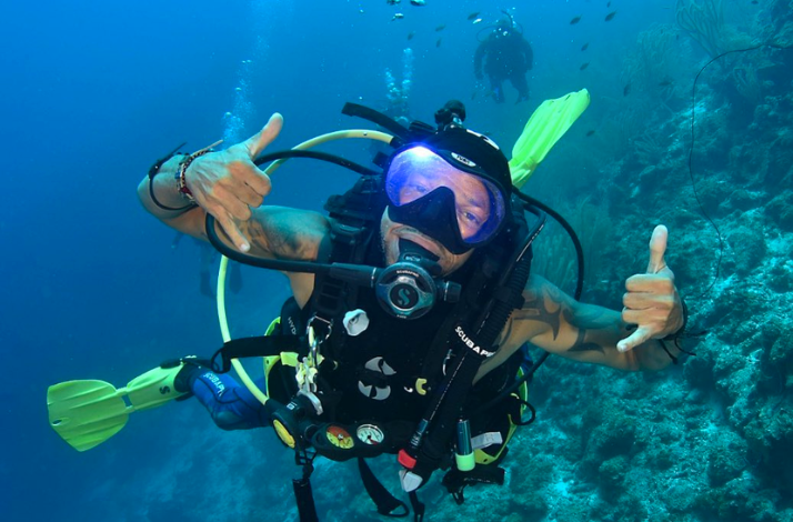 A scuba diver smiles at the camera, giving a 