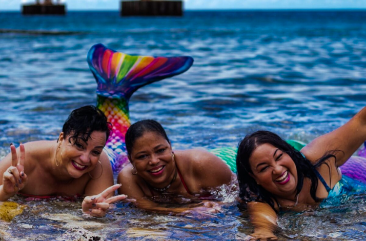 Three people in vibrant mermaid tails pose playfully together in the shallow ocean water
