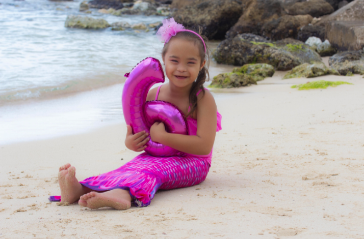 A child in a pink mermaid costume sits on the sand, holding a pink number balloon