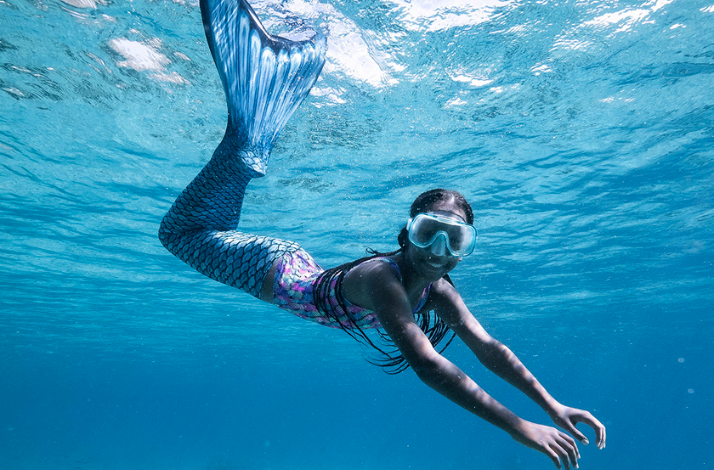 A person in a blue mermaid tail swims underwater in clear, tropical ocean