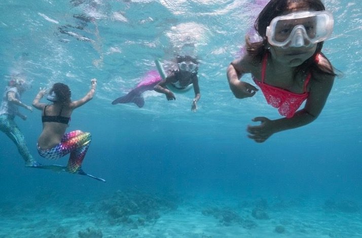 A group of girls wearing mermaid tails and goggles swims together in the clear, shallow ocean water.