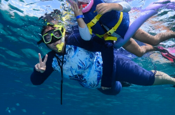Two snorkelers, one using a pool noodle, swim together in clear blue water, making peace signs.