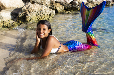 A person in a colorful mermaid tail lies on the shallow shoreline near rocky beach formations