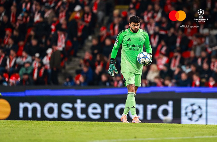 Arsenal goalkeeper holding the ball in front of a packed stadium during a UEFA Champions League 2025/26 match at SK Slavia Praha, Upper-right corner: Mastercard logo + UEFA Champions League logo with text 'official sponsor'.