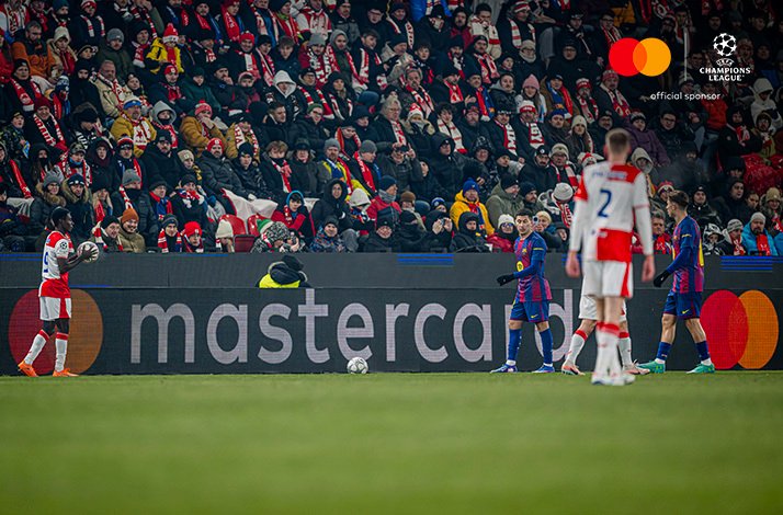 Players from SK Slavia Praha and FC Barcelona competing on the pitch. Upper-right corner: Mastercard logo + UEFA Champions League 'official sponsor' logo.