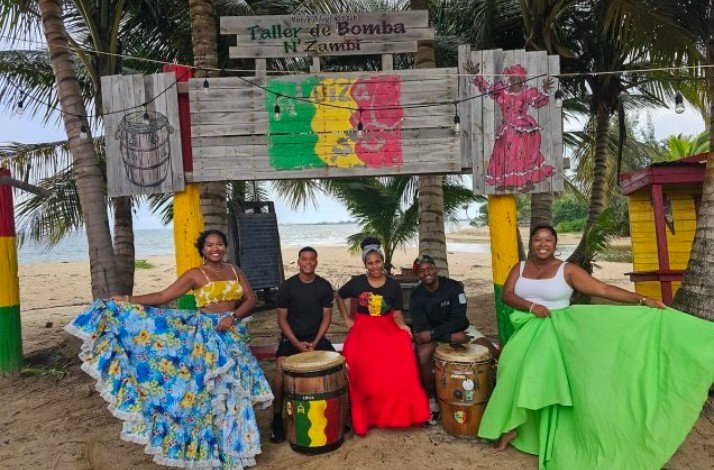 Bomba dancers wearing eye–catching traditional dresses posing next to drummers on a beach.
