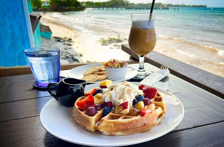 A delicious waffle with ice cream and fresh fruits served with an iced coffee on the table with beach view