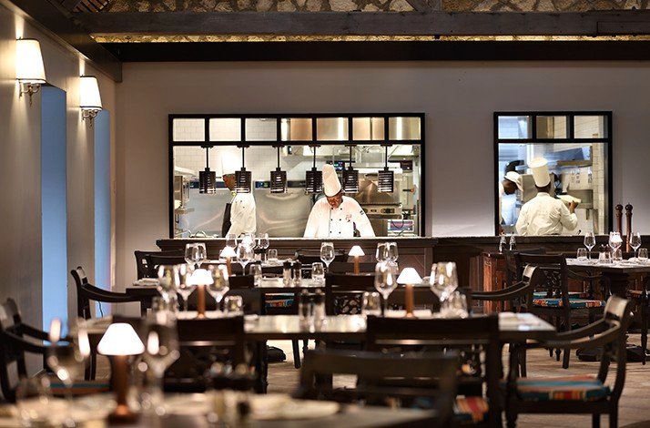 Indoor of Half Moon resort, a group of tables set up waiting for commensals, and chefs at the background working at the kitchen