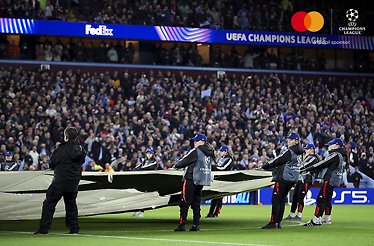 Staff unfolding a pitch cover across a stadium, Mastercard and UEFA Champions League 'official sponsor' logos in the upper-right corner.