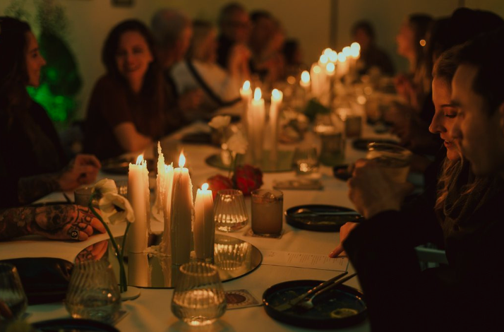 Long communal dinner table lit by tall candles as guests chat and dine in warm light.