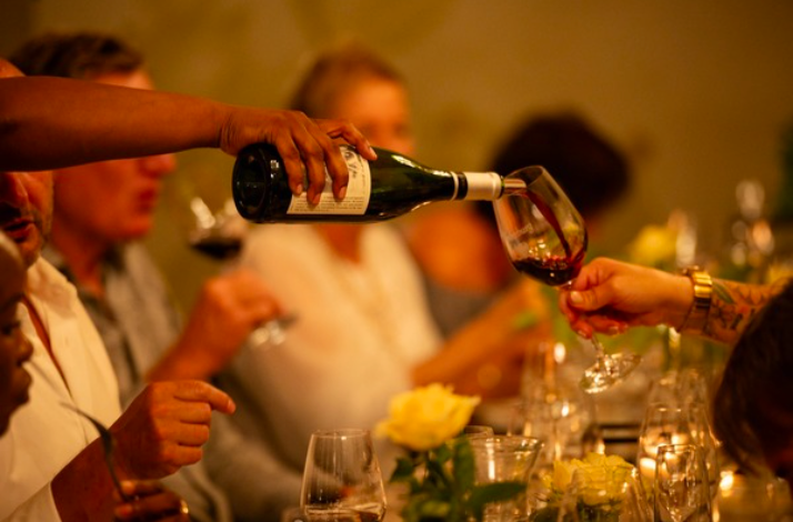 Server pouring red wine into a glass at a crowded dinner table.