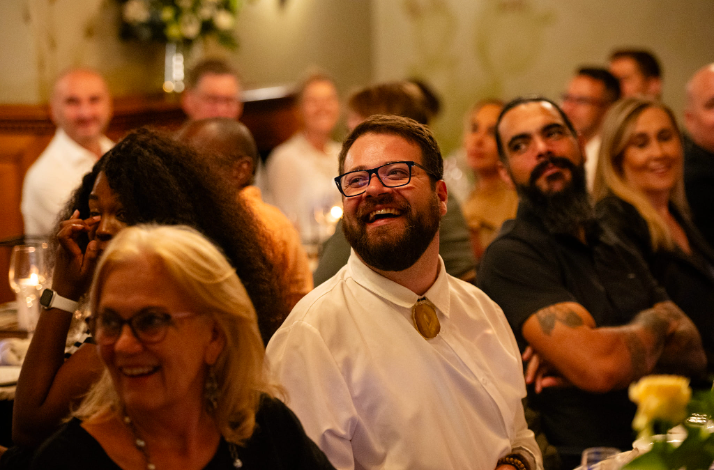 Group of dinner guests seated closely together, smiling and looking toward a speaker