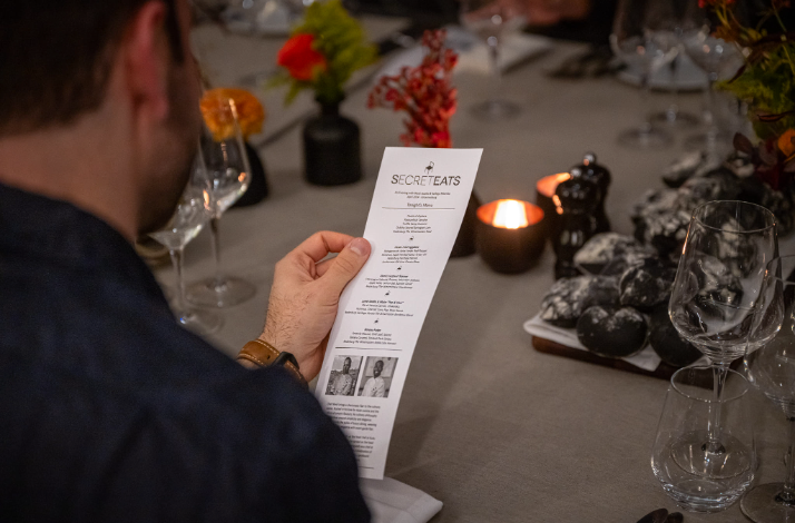 Guest holding a printed Secret Eats menu at a candlelit dinner table set with wine glasses.