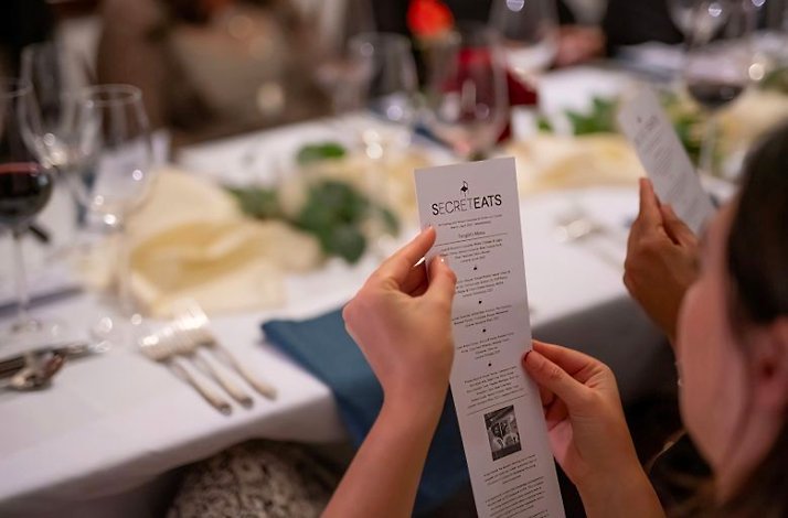 A guest reading the printed SecretEATS menu card at a table set with greenery and glassware