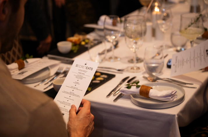 A guest holding a SecretEats menu at an elegantly set dinner table with fine glassware.