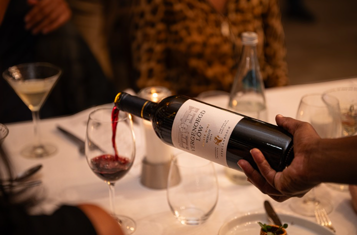 Red wine from a Vergenoegd Löw Merlot bottle being carefully poured into a glass at the table.