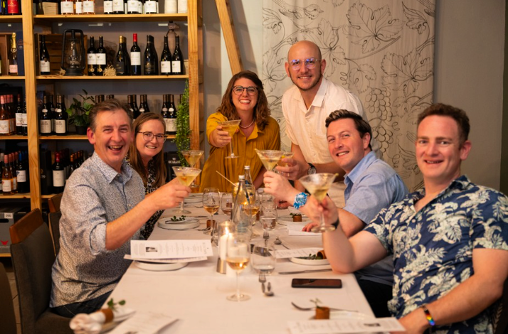 Six happy guests raising martini glasses for a celebratory toast in a cozy wine-cellar venue.