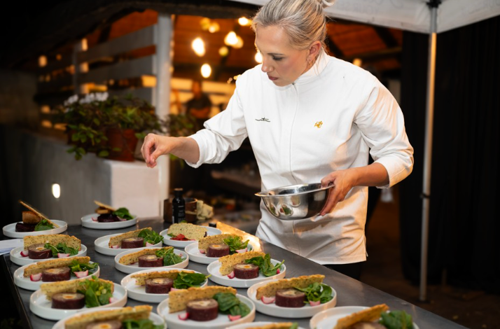 A chef in a white uniform plating gourmet meat terrines with fresh greens on a long prep counter.