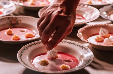 Hand placing a small dollop of cream onto plated pink dessert soup with garnishes.