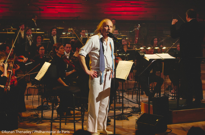 Philippe Katerine singing at a microphone stand on stage at Philharmonie de Paris, with a conductor and full orchestra performing behind him. ©Ronan Thenadey / Philharmonie de Paris.