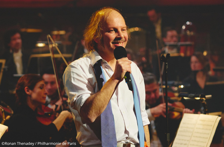 Philippe Katerine smiling and singing into a handheld microphone, with orchestra musicians visible in the background. ©Ronan Thenadey / Philharmonie de Paris.