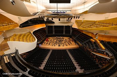Philharmonie de Paris concert hall viewed from above, featuring curved architecture, amber seating tiers, and orchestra on stage. Crédit Gil Lefauconnier.