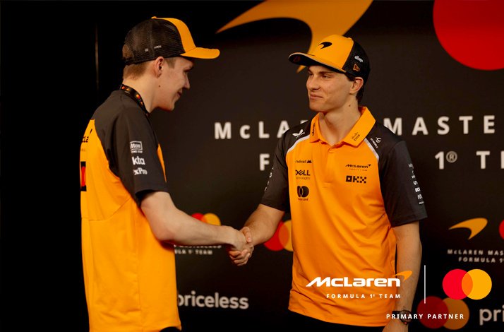 Two people wearing McLaren F1 merchandise in front of a McLaren in collaboration with Mastercard Banner.