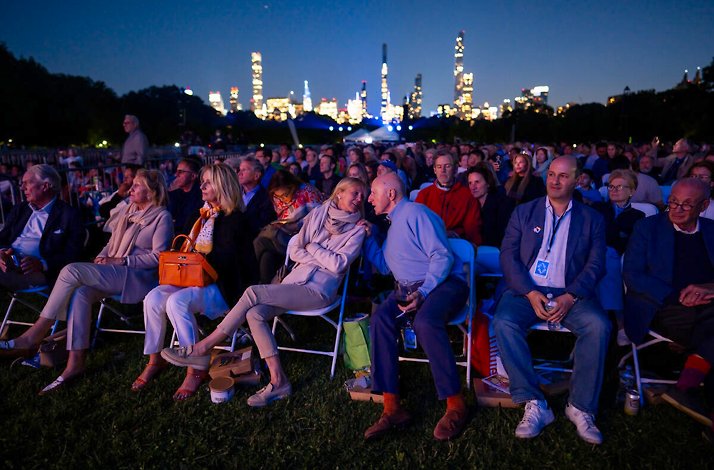 An outdoor audience watches the New York Philharmonic Concerts in the Parks in Central Park against the NYC skyline.