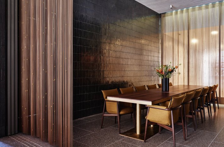A dining area features a wooden table with mustard-colored chairs, set against a wall of dark-brown, rectangular tiles. 