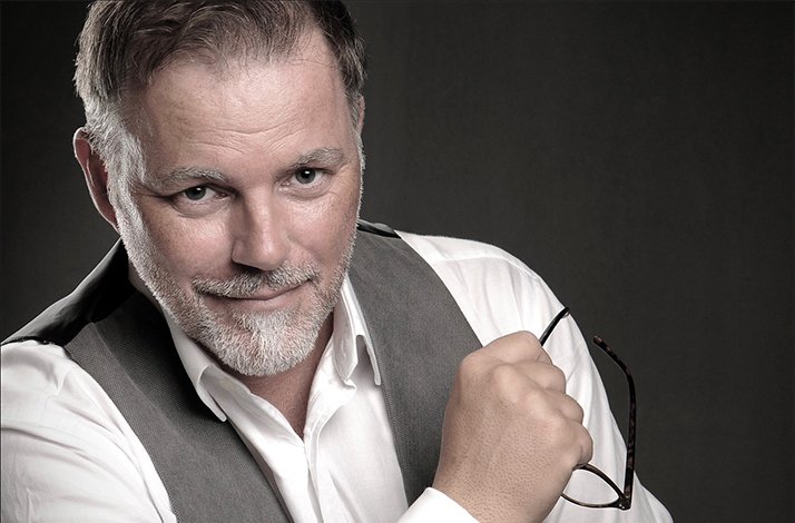 Jean-Peer Krause, dressed in a white shirt and grey vest, holding eyeglasses against a dark background.