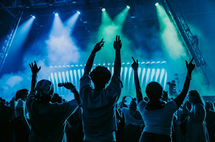 Silhouetted crowd members cheering with arms raised at AO Live Presents, surrounded by teal stage lighting and atmospheric smoke effects.