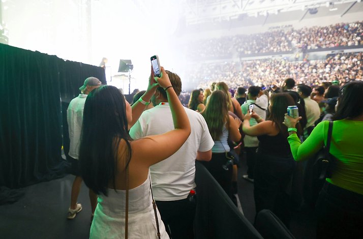 Side-of-stage view at John Cain Arena, with a Mastercard cardholder filming AO Live Presents while crowds fill the arena in the background.