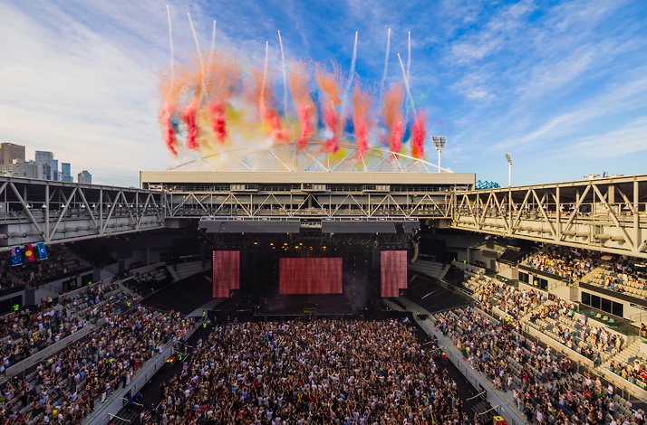 Colourful confetti cannons bursting above John Cain Arena at AO Live Presents, with a packed crowd on the floor and Melbourne skyline beyond.